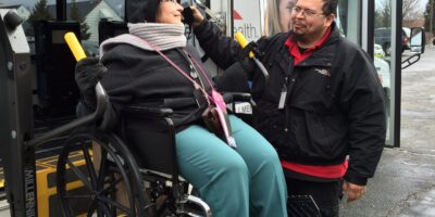 Smiling woman in a wheelchair exiting a paratransit bus via lift, being helped by a transit worker