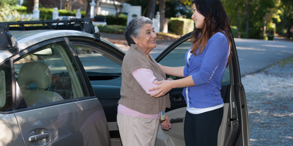 Photo of a young woman assisting an older woman beside a car.
