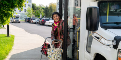 Smiling woman with a walker and bag exiting a paratransit bus