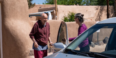 Volunteer driver helping older man approaching car with pueblos style building in background