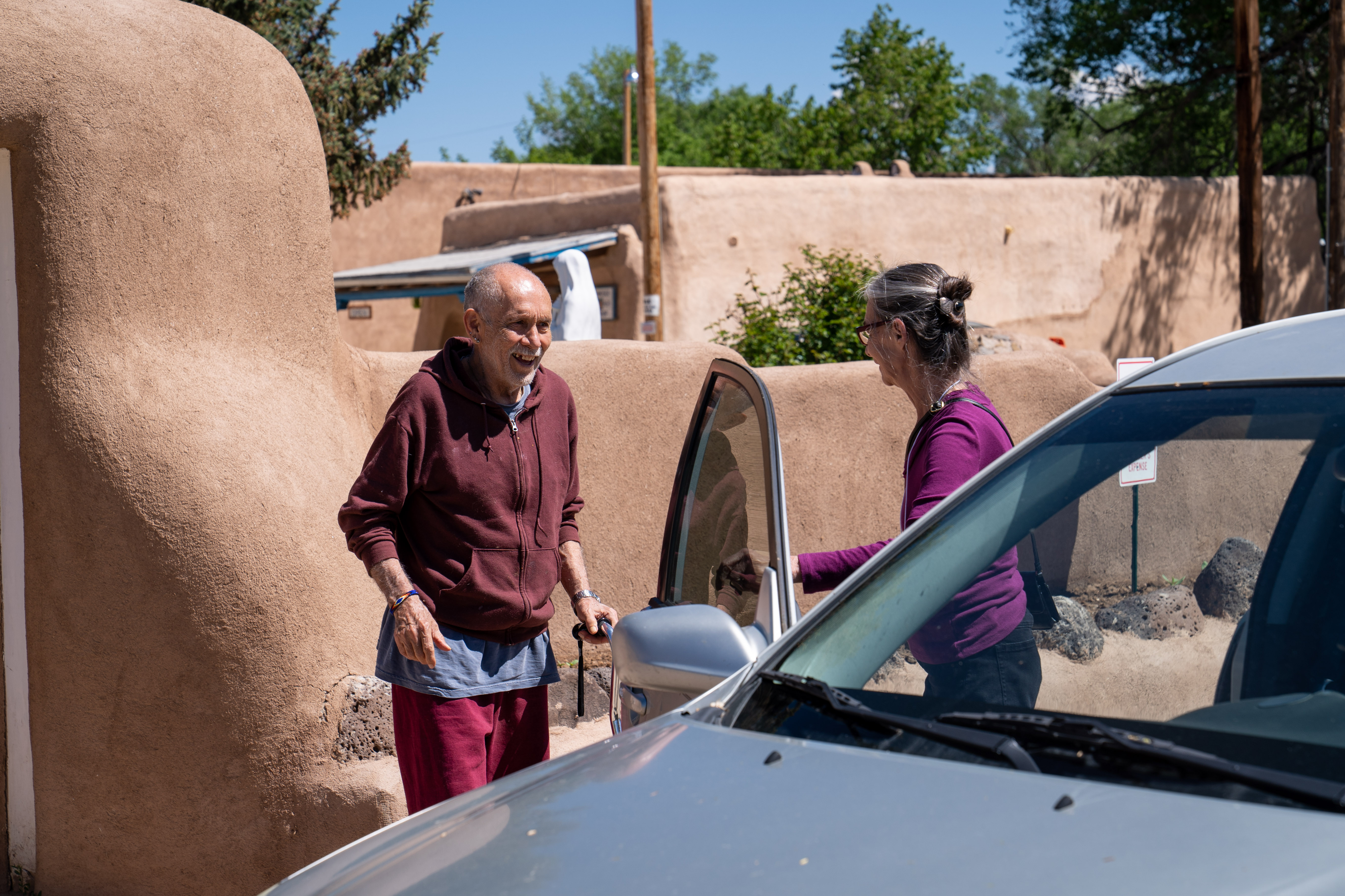 Volunteer driver helping older man approaching car with pueblos style building in background