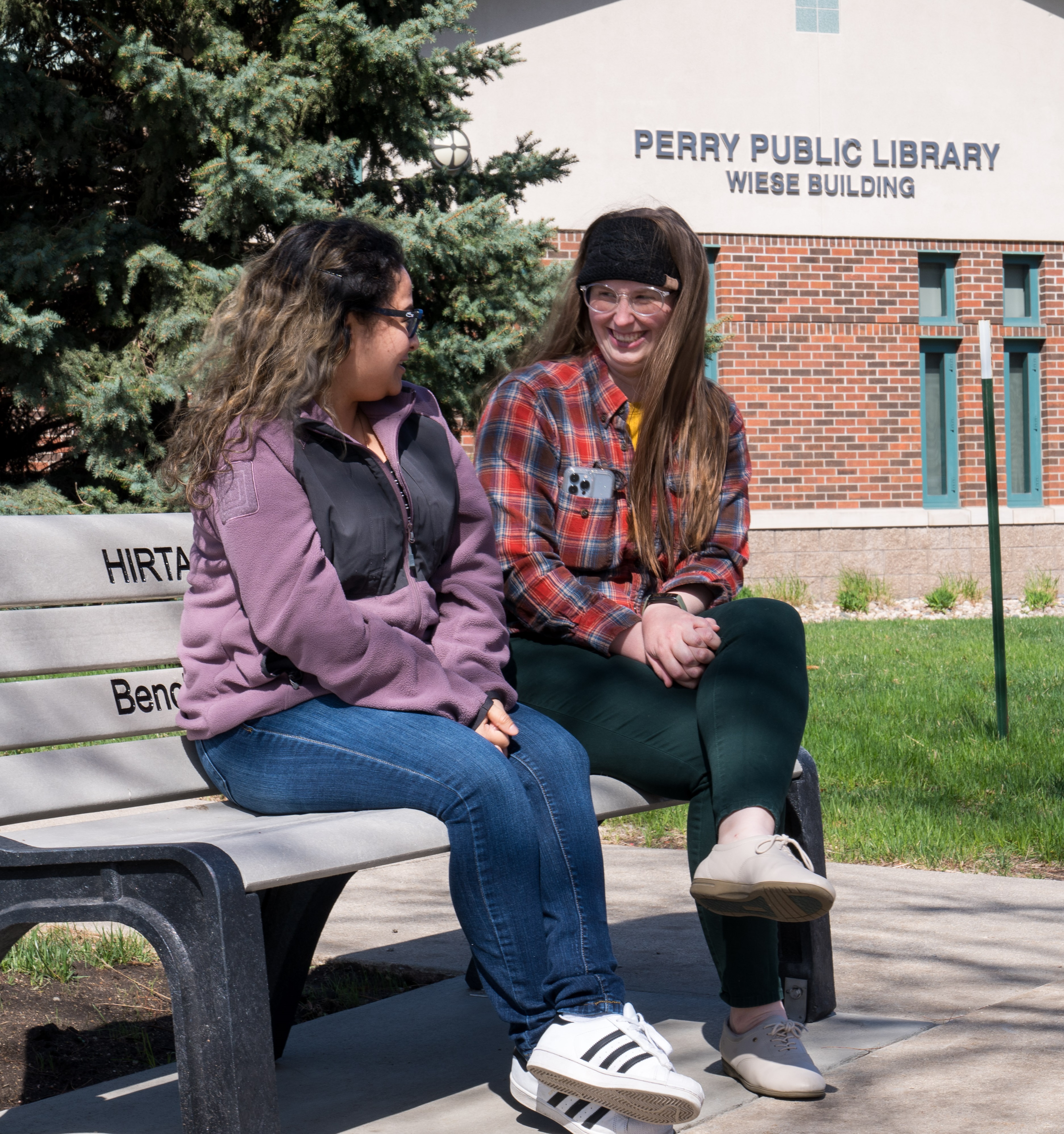 Two women sitting on bus stop bench in front of Perry Public Library
