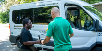 Visually impaired man with cane being helped into van by man in green shirt