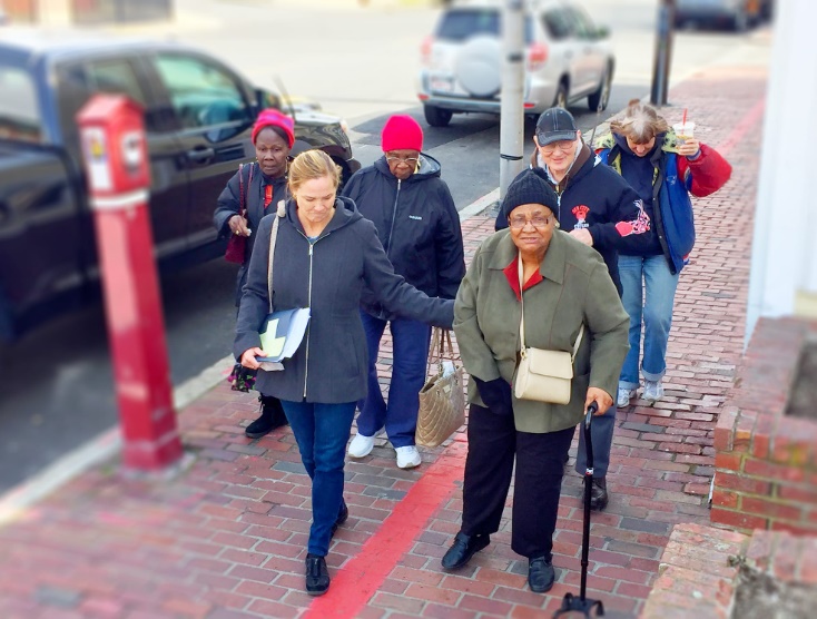 A woman helping an older woman with a can walk on a sidewalk with other people walking behind them.