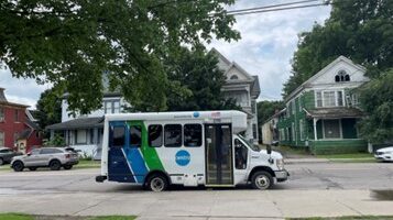 White bus with blue and green stripes on a street in front of houses.
