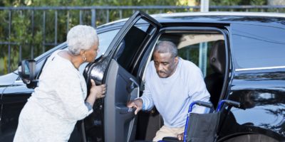 A senior African American man is getting out of a car, into his wheelchair. His wife is helping him, holding the door open.