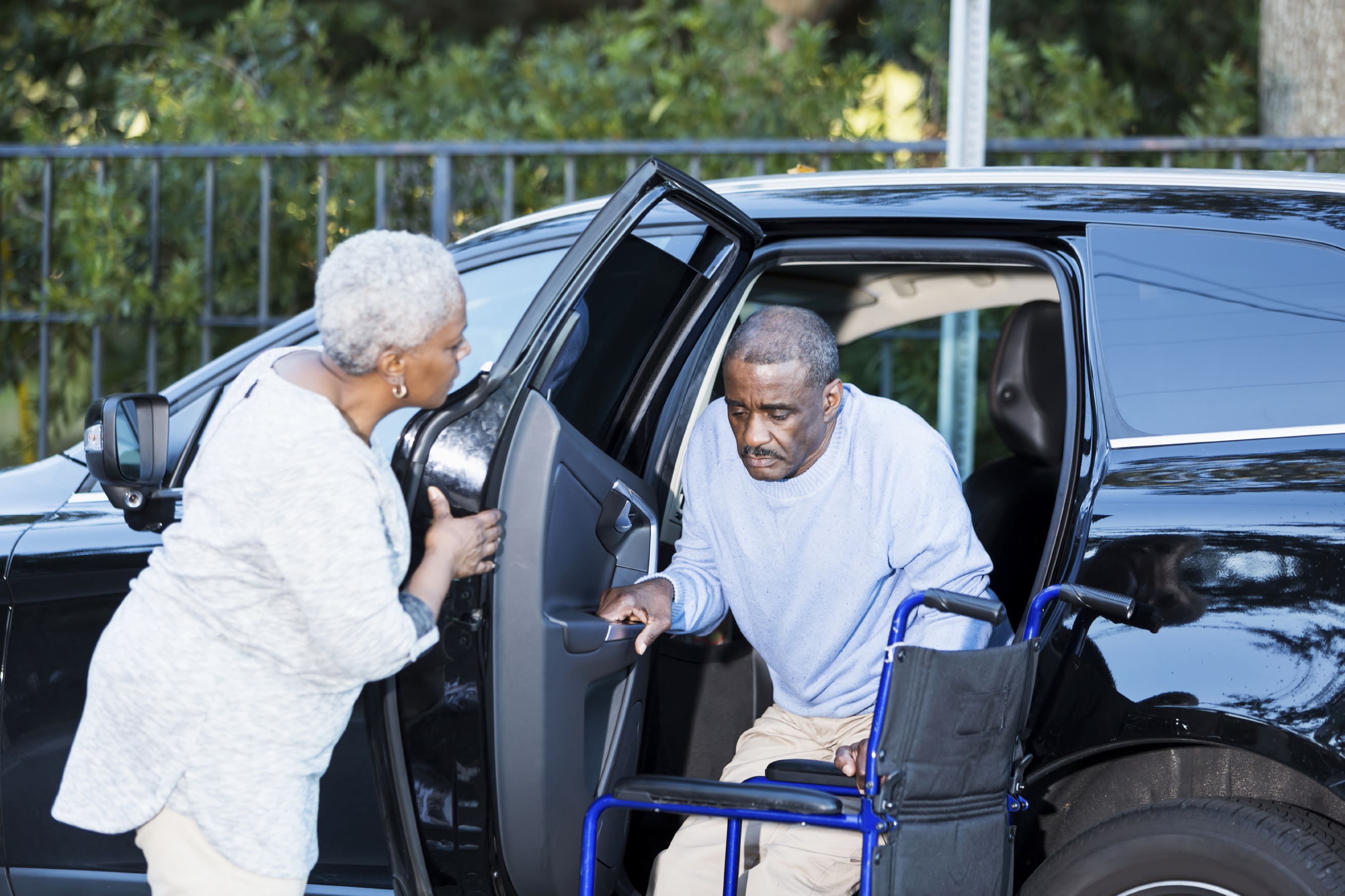 A senior African American man is getting out of a car, into his wheelchair. His wife is helping him, holding the door open.