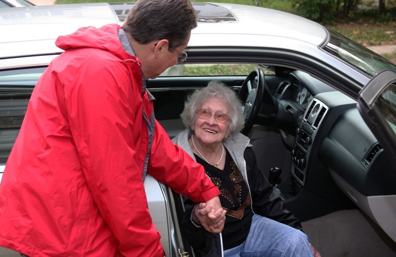 man in red jacket helping older woman out of passenger seat of car