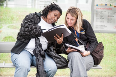 Two women sitting at a bus stop, reading a bus schedule. 